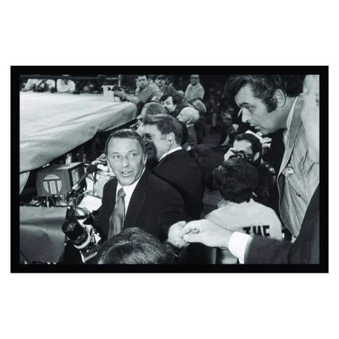 Black and white photo of a smiling man at a boxing event surrounded by celebrities and photographers.